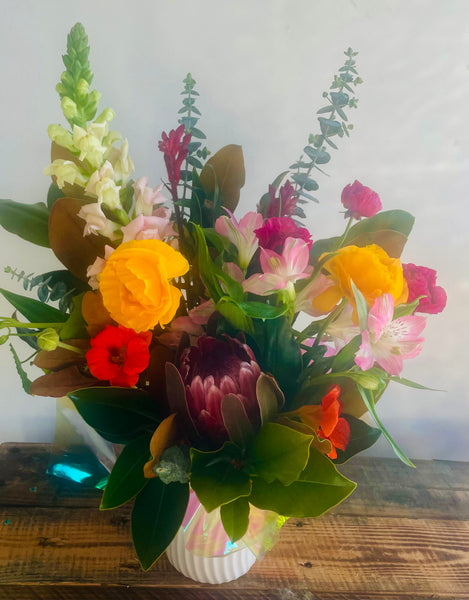 A vibrant mixed floral arrangement in a white ribbed vase, featuring yellow roses, pink alstroemeria, red snapdragons, a dark purple protea, and green foliage, displayed on a rustic wooden surface against a plain light background.