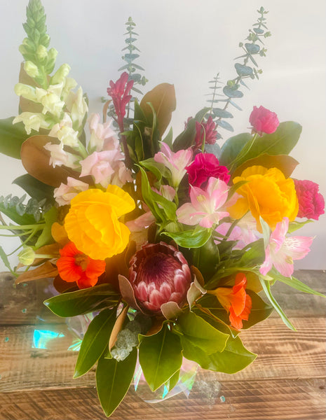 A vibrant mixed floral arrangement featuring a protea, yellow roses, pink and white alstroemeria, orange flowers, and green foliage, displayed in a clear glass vase on a rustic wooden surface.